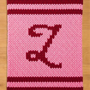 Puede incluir: Una manta de punto rosa y burdeos con la letra "I" en burdeos. La manta tiene un aspecto grueso y texturizado, con la letra centrada en el campo rosa. Rayas burdeos bordean la parte superior e inferior.