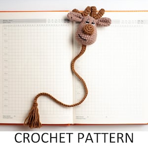 May include: A brown crocheted reindeer bookmark with a tassel. The bookmark is laying open on a white background with a blank orange planner.