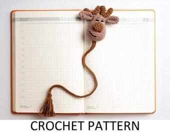 May include: A brown crocheted reindeer bookmark with a tassel. The bookmark is laying open on a white background with a blank orange planner.