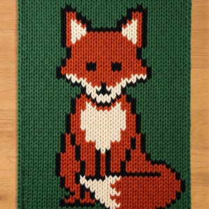 May include: A knitted square rug with a pixelated fox design. The fox is mainly orange, with white and black details, set against a green background. The rug has a thick, textured appearance.