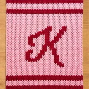 Könnte beinhalten: Ein gestricktes Quadrat in Rot- und Rosatönen. Die oberen und unteren Ränder sind tiefrot, mit einem helleren roten Streifen in der Mitte. In der Mitte befindet sich ein großes, stilisiertes "A" in dunklerem Rot.
