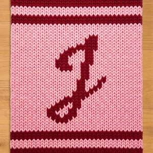 Könnte beinhalten: Ein gestrickter, rechteckiger Artikel mit rosa Hintergrund und einem weinroten Buchstaben "J". Die oberen und unteren Ränder sind weinrot, mit zwei weinroten Streifen. Der Artikel ist wahrscheinlich eine Decke oder ein Kissen.