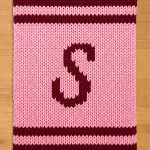 Puede incluir: Una manta rosa tejida con un borde y rayas burdeos. Una gran letra "S" burdeos está centrada en la manta. La manta está hecha de un hilo grueso y texturizado.
