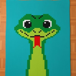 May include: A turquoise blanket with a pixelated green snake design. The snake has large black eyes, a red tongue, and a light green body. The blanket is made of a soft, knitted material.