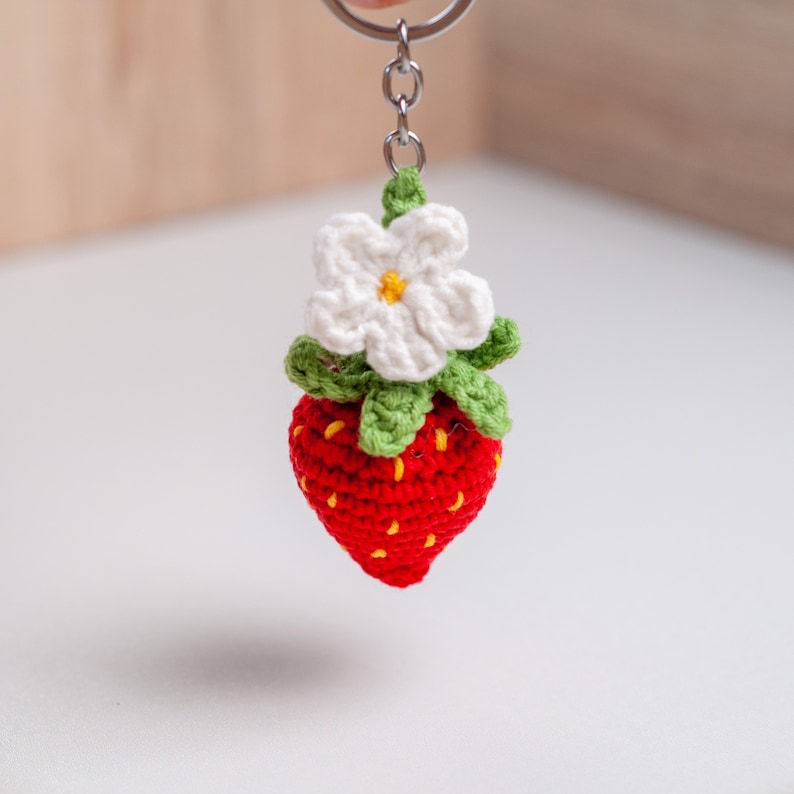 Puede incluir: Un llavero de fresa tejido a crochet con una flor blanca en la parte superior. La fresa es roja con semillas amarillas y hojas verdes.