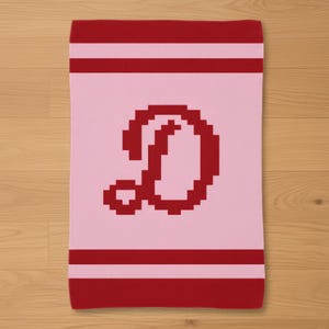 May include: A pink and red knit blanket featuring a large, pixelated letter "D" in red. The blanket has red stripes at the top and bottom, with a lighter pink stripe in between.