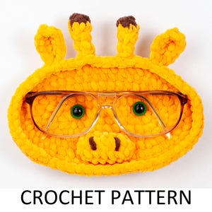 May include: A crocheted giraffe-shaped glasses holder with brown horns and green eyes. The holder is made of yellow yarn and has a clear plastic frame for holding glasses.