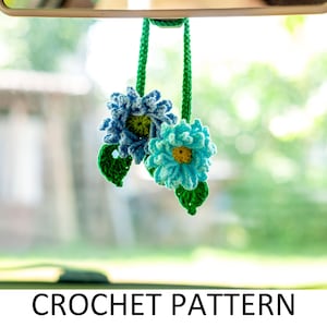 以下が含まれることがあります： リアビューミラーに吊るされた、緑の葉が付いた青い花2つのためのクロシェパターン。花は青と白の糸で編まれ、葉は緑の糸で編まれています。