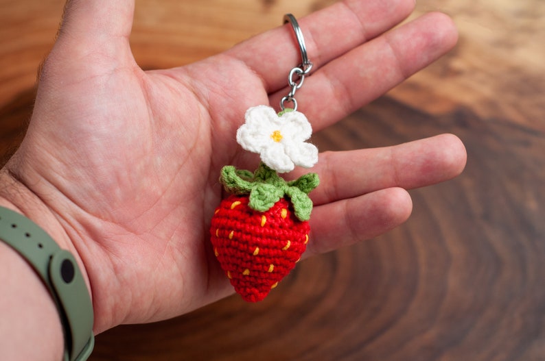 Puede incluir: Un llavero de fresa tejido a crochet con una flor blanca y hojas verdes. El llavero est&aacute; unido a un anillo de metal plateado.