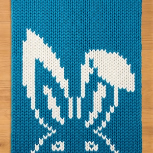 May include: A square, knitted blue pillow with a white bunny face design. The pillow is made of thick yarn and has a textured appearance. The bunny face is centered on the pillow.
