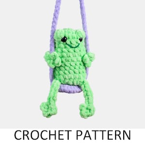 May include: A green crocheted frog toy sitting on a purple crocheted swing. The frog has a smiling face with black eyes and a green mouth.