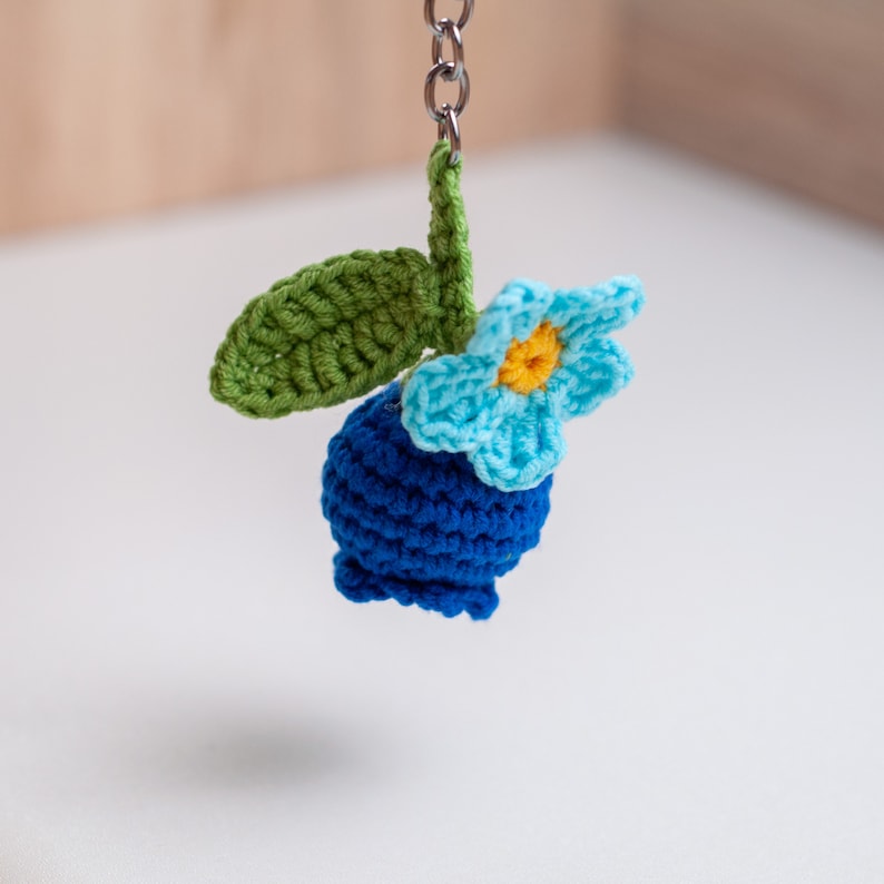 Op de afbeelding: Een gehaakte blauwe bessenbedel met een blauwe bloem en een groen blad. De bedel is bevestigd aan een zilveren sleutelhanger.