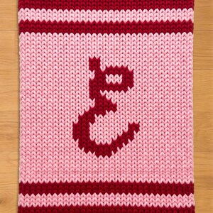 May include: A knitted blanket in shades of pink and burgundy. The blanket features a large, stylized letter "E" in burgundy in the center. The top and bottom edges, along with a stripe, are burgundy. The background is light pink.