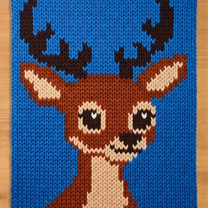 May include: A knitted square featuring a cartoon deer with brown antlers, a brown body, and a white chest. The deer has large black eyes and a black nose. The background is a solid blue color.