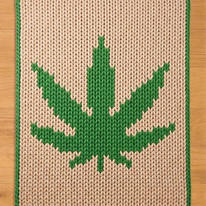 May include: A rectangular, knitted rug featuring a large green cannabis leaf design on a cream-colored background. The leaf is centrally placed, with a green border outlining the rug's edges. The rug is made of thick yarn.