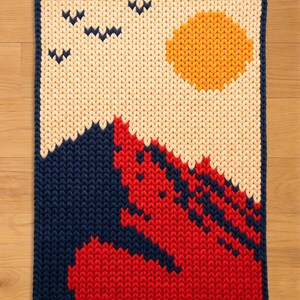 May include: A knitted wall hanging with a landscape design. The artwork features a blue mountain range, a red and blue foreground, a yellow sun, and three blue birds against a cream-colored background. The edges are trimmed with a dark blue border.