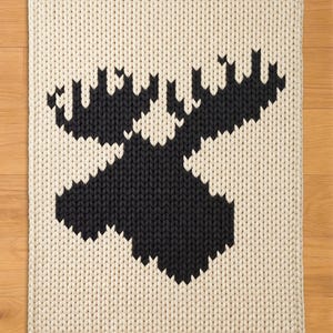 Könnte beinhalten: Cremefarbene Strickdecke mit einer großen schwarzen Elchsilhouette. Die Decke hat ein dickes, strukturiertes Aussehen mit einem detaillierten, geometrischen Design des Elchkopfes und Geweihs. Die Kanten sind sauber mit einem Strickrand versehen.