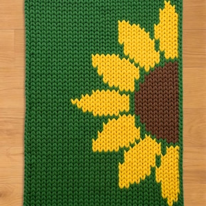 May include: A knitted green rectangular rug with a large sunflower design. The sunflower has yellow petals and a brown center. The rug is made of thick yarn and has a textured appearance.