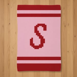 May include: A pink blanket with a large red letter "S" in the center. The blanket has red stripes at the top and bottom. The blanket is displayed on a wooden surface.