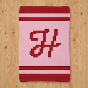Puede incluir: Una manta rosa con borde y rayas rojas. La manta presenta una gran letra "H" roja pixelada en el centro. El diseño es simple y moderno, adecuado para una guardería o dormitorio.