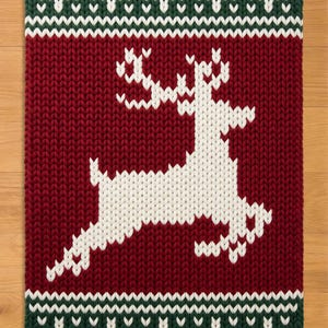 May include: A rectangular knit blanket featuring a large white reindeer silhouette centered on a deep red background. The top and bottom borders consist of a dark green and white geometric chevron pattern, all crafted with a chunky knit texture.