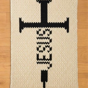 May include: Cream-colored, knitted rectangular rug with a large black syringe design. The syringe features a cross-shaped top and a detailed body, creating a bold, graphic statement. The rug's texture is visible, adding depth to the design.