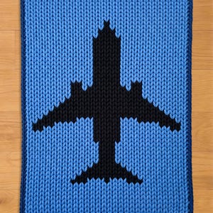 May include: A knitted rug with a blue and black airplane design. The rug has a light blue background, a dark blue border, and a black airplane in the centre. The rug is rectangular.