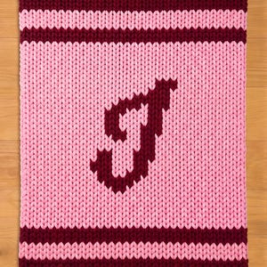 Può includere: Una coperta lavorata a maglia in rosa e bordeaux. La coperta presenta una grande lettera "J" bordeaux al centro su uno sfondo rosa. Strisce bordeaux bordano i bordi superiore e inferiore, creando un'estetica accogliente e fatta a mano.