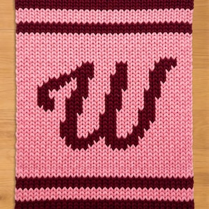 Può includere: Una coperta lavorata a maglia rosa e bordeaux con le lettere "TW" in bordeaux. La coperta ha un bordo a righe in bordeaux e rosa. Il design a maglia grossa conferisce alla coperta un aspetto accogliente e fatto a mano.