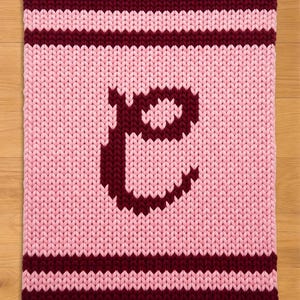 Könnte beinhalten: Eine rechteckige Strickdecke in Rosa- und Burgundertönen. Die Decke zeigt einen großen Buchstaben "E" in Burgunderrot in der Mitte. Burgunderfarbene Streifen säumen die oberen und unteren Kanten, mit einem rosa-burgunderfarbenen Streifenmuster.