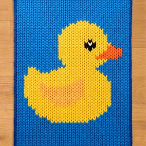 Puede incluir: Una alfombra rectangular azul tejida con un diseño de pato amarillo. El pato tiene un pico naranja, ojos negros y un cuerpo amarillo con un centro naranja. La alfombra tiene un borde azul y está hecha de material tejido.