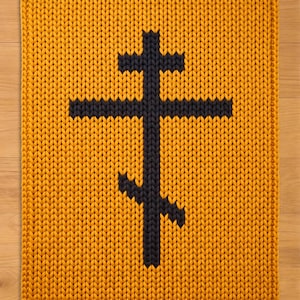 May include: A rectangular, knitted rug in a mustard yellow color. A black, Eastern Orthodox cross is centered on the rug. The rug has a thick, textured appearance, with visible knit stitches.
