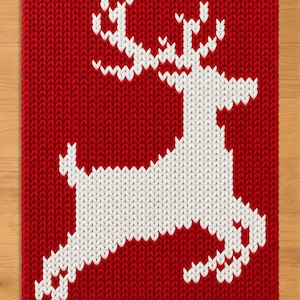 May include: A rectangular knit textile featuring a bold red background with a large, white, stylized reindeer silhouette centered in the middle. The knit pattern is consistent throughout, showing a clear, textured stitch design in both colors.
