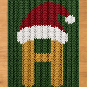 May include: A rectangular knit textile featuring a dark green background with a large, golden-yellow letter H centered. The letter is topped with a red and white knit Santa hat, complete with a white pom-pom detail on the right side.