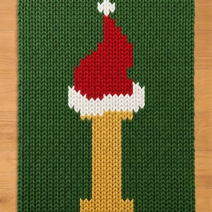 Puede incluir: Un tejido rectangular de punto verde que presenta una letra I amarilla central rematada con un gorro de Papá Noel rojo y blanco. El patrón de punto es consistente en toda la pieza, mostrando un contraste claro entre el fondo verde y la letra.