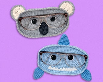 May include: Two crocheted eyeglass holders, one shaped like a koala and the other shaped like a shark. Both holders are light blue and gray and have brown glasses resting on them.