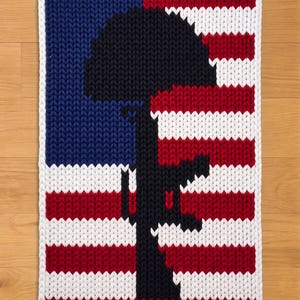 Op de afbeelding: Een gebreide rechthoekige deken met een zwart silhouet van een soldatenmonument tegen een achtergrond van de Amerikaanse vlag. Het vlagontwerp bevat blauwe, rode en witte strepen en een witte rand.