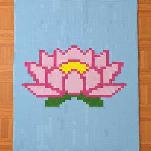 May include: A light blue knitted blanket featuring a pixelated pink lotus flower with a yellow center and green leaves. The blanket is rectangular and appears to be handmade.