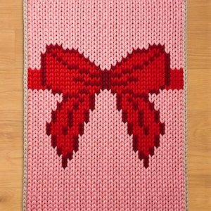 Può includere: Tappeto lavorato a maglia con un grande motivo a fiocco rosso su sfondo rosa. Il fiocco è di una tonalità di rosso più scura e il tappeto ha un bordo beige chiaro. Il tappeto è rettangolare e sembra fatto a mano.