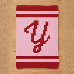 May include: A rectangular pink blanket with a red letter "Y" in the center. The letter is pixelated. The top and bottom edges have red stripes. The blanket is on a wooden surface.