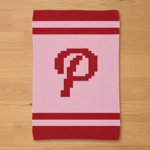 May include: A pink knitted scarf featuring a large red "P" in the centre. The top and bottom edges have red stripes. The scarf is rectangular and appears to be made of soft material.