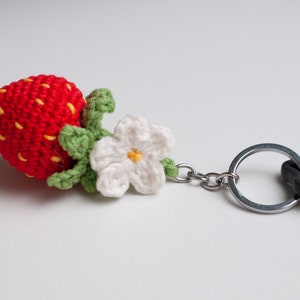 Puede incluir: Un llavero de fresa tejido a crochet con un tallo y hojas verdes, una flor blanca con un centro amarillo y un llavero plateado.