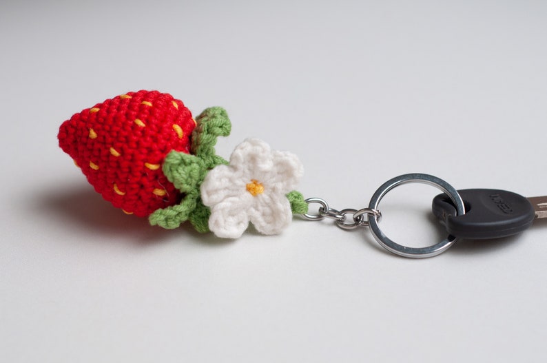 Puede incluir: Un llavero de fresa tejido a crochet con un tallo y hojas verdes, una flor blanca con un centro amarillo y un llavero plateado.