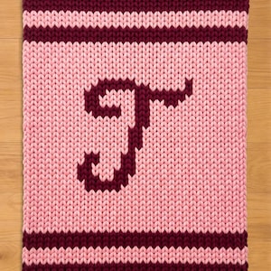 May include: A knitted rectangular item with a light pink background and a dark burgundy letter "T". The top and bottom borders are dark burgundy, with a light pink stripe in between. The item is made of thick yarn.