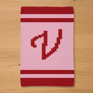 Puede incluir: Bufanda rosa rectangular con una letra "V" roja en el centro. La bufanda tiene rayas rojas en la parte superior e inferior. La bufanda está hecha de un material de punto.