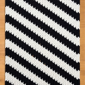 Pode incluir: Um tapete de malha preto e branco com um padrão arrojado de riscas diagonais. O tapete é retangular e apresenta riscas espessas e texturizadas. As cores contrastantes criam um efeito visual marcante, adequado para decoração de interiores.