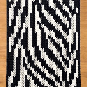 May include: A rectangular, hand-knitted rug with a bold, geometric pattern. The design features alternating bands of black and white, creating a striking visual contrast. The rug's edges are neatly finished, and the overall aesthetic is modern and graphic.