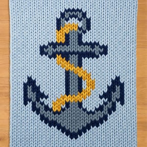 May include: A light blue chunky knit blanket featuring a large, centred anchor design. The anchor is rendered in shades of dark navy blue, grey, and white, with a prominent yellow rope wrapped around the central shaft of the anchor structure.