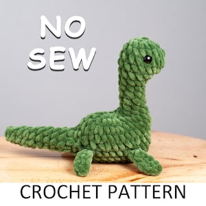 Puede incluir: Un juguete de dinosaurio de crochet verde con ojos negros. El juguete está sentado sobre una superficie de madera. El texto "NO SEW CROCHET PATTERN" está escrito debajo del juguete.