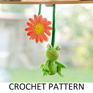 May include: Crochet pattern for a green frog and a pink flower hanging from a car mirror. The frog has a smiling face and is crocheted in a light green yarn. The flower is crocheted in a pink yarn with a yellow center.